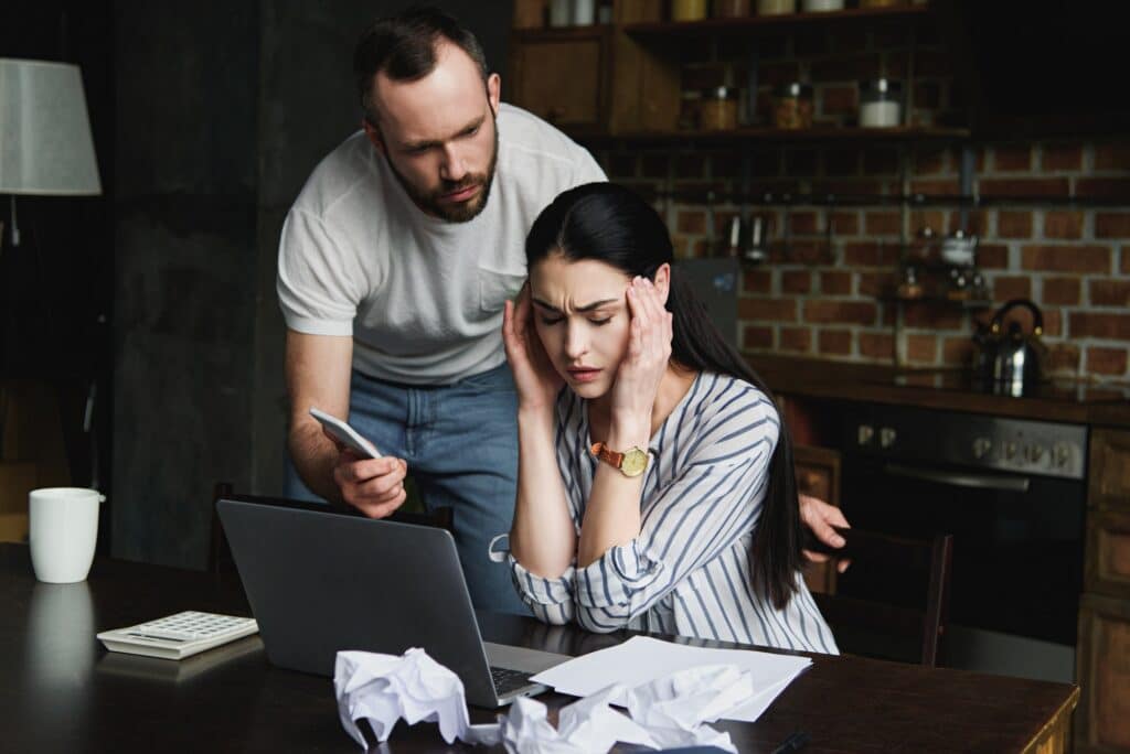 stressed young woman sitting at table with laptop and crumpled papers while her angry husband